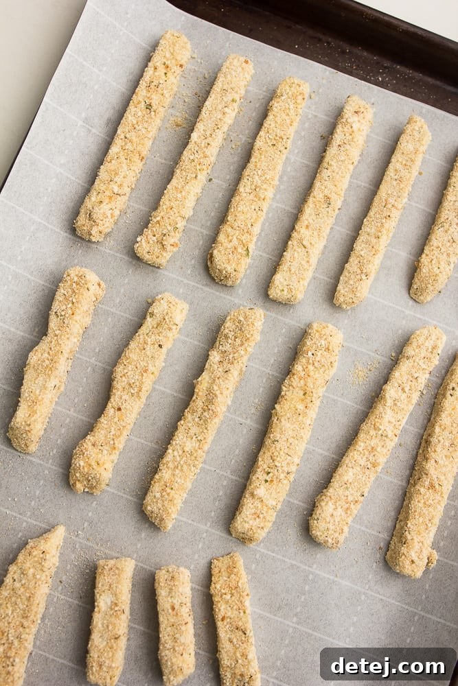 battered and breaded sticks of vegan cheese on a baking sheet with parchment paper, ready for freezing