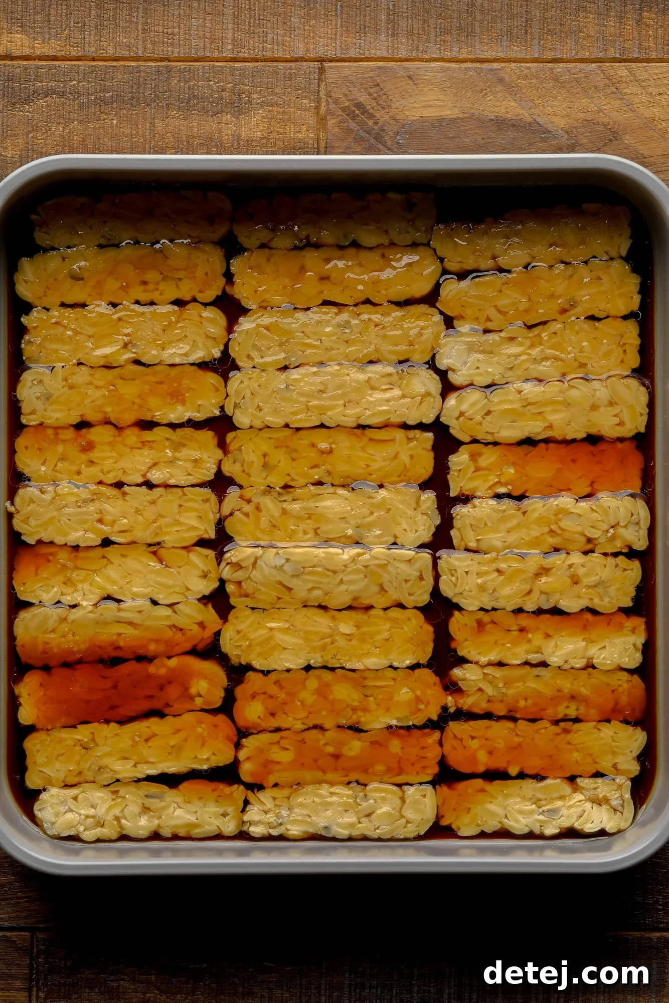 Thinly sliced tempeh soaking in a rich, brown marinade in a shallow dish, ready for refrigeration.