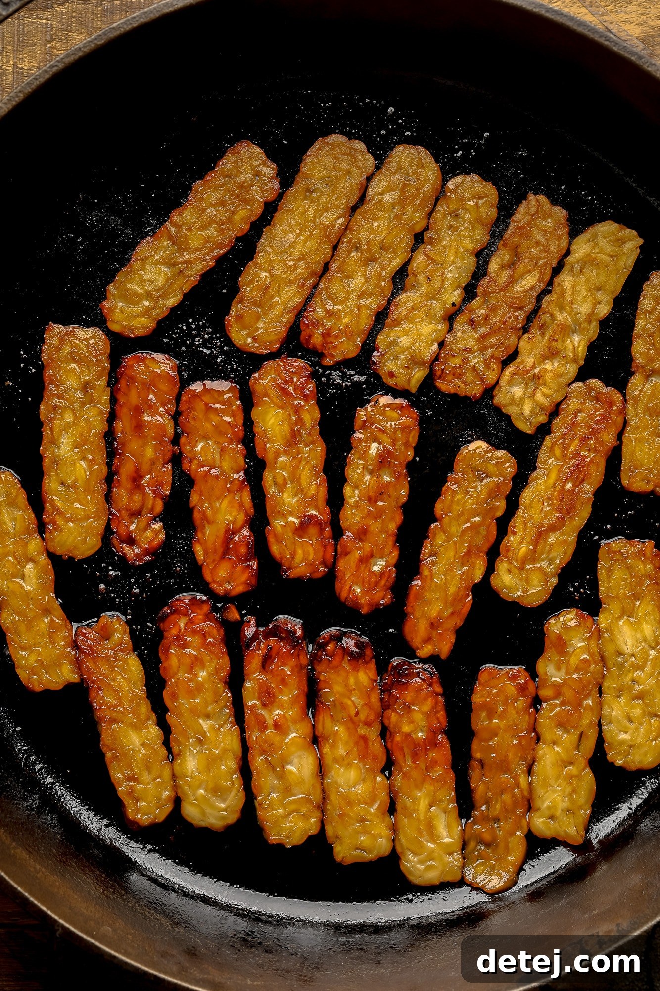 Slices of marinated tempeh bacon sizzling and turning golden brown in a hot black skillet, perfectly pan-fried.