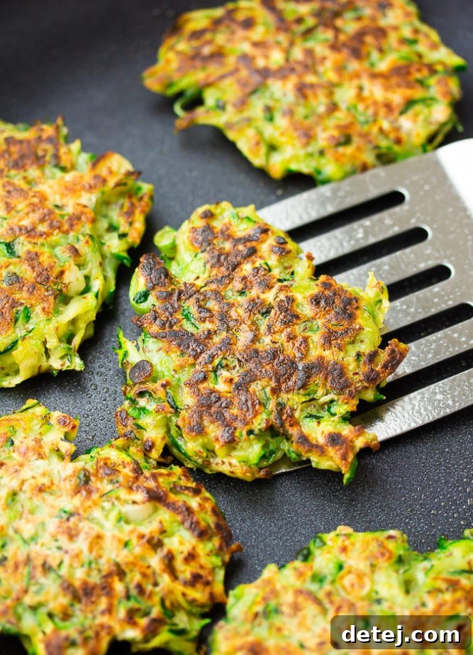 A frying pan filled with golden-brown vegan zucchini fritters, with one being lifted out by a spatula, showcasing its crisp exterior.