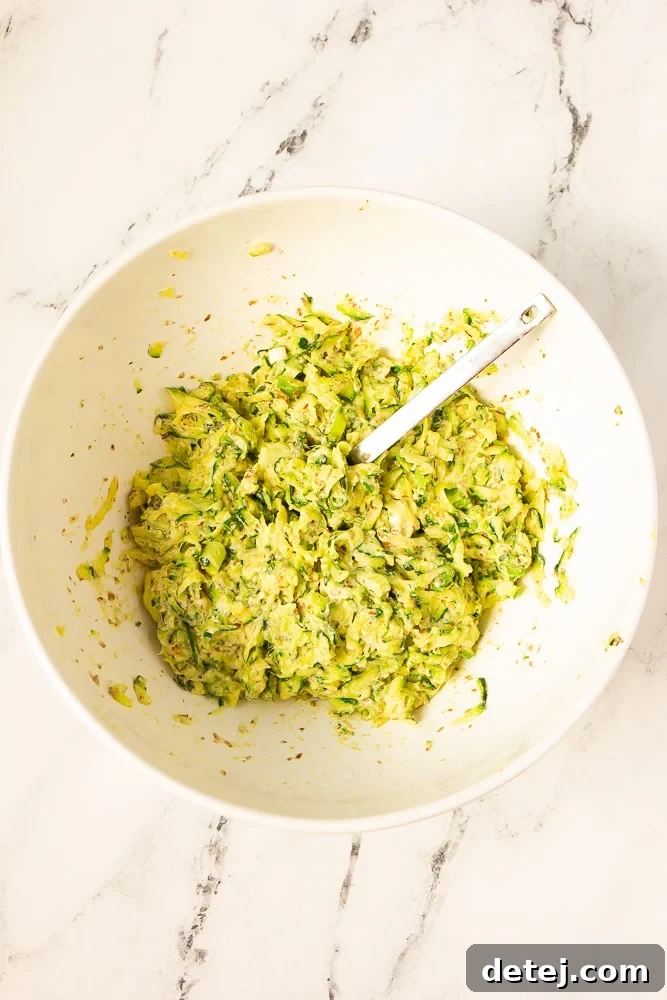 A pristine white bowl filled with the vibrant green zucchini fritter mixture, with a spoon resting inside, ready for cooking.