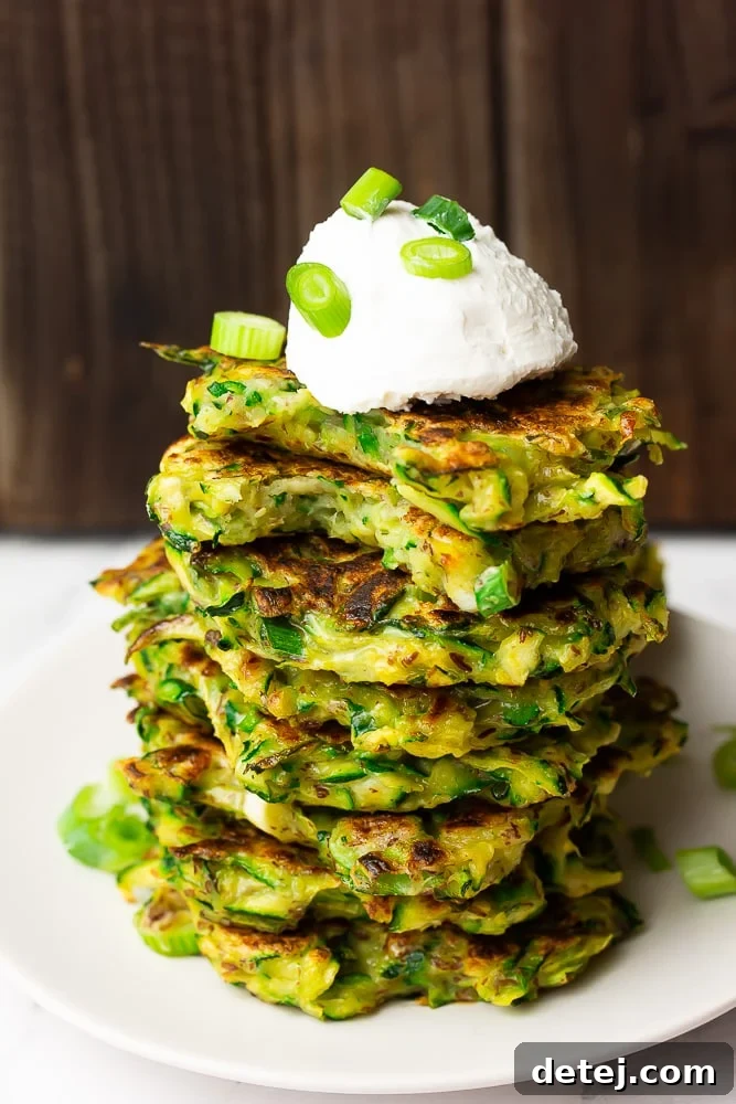 A close-up of a stack of vibrant green zucchini fritters, topped with a dollop of creamy vegan sour cream and a sprinkle of fresh chives.