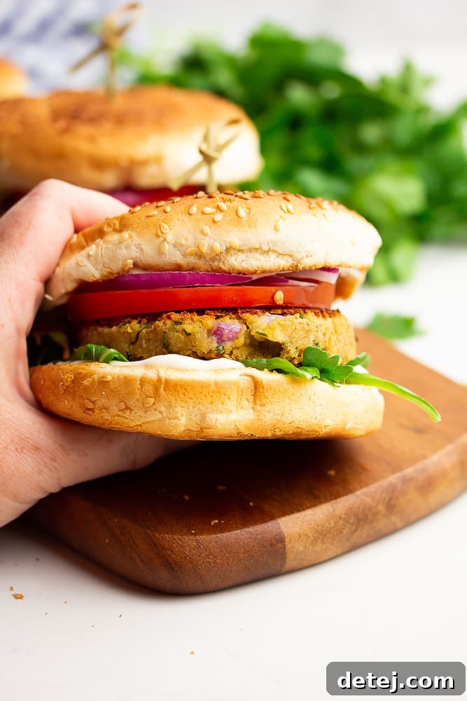 Hand holding a perfectly cooked chickpea burger with fresh toppings, with more burgers in the background, ready to be served.