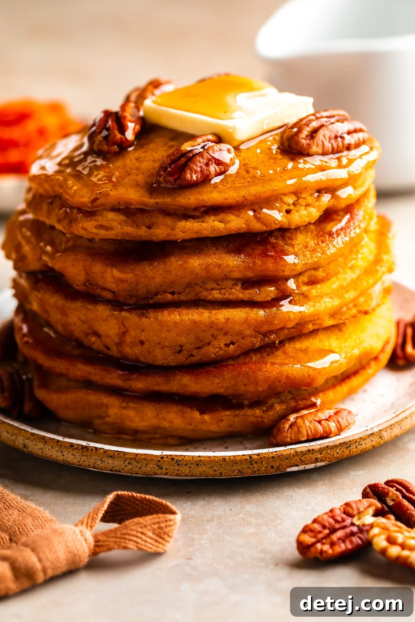 Close-up on a stack of golden vegan pumpkin pancakes, lavishly topped with flowing maple syrup, scattered pecans, and a pat of vegan butter.