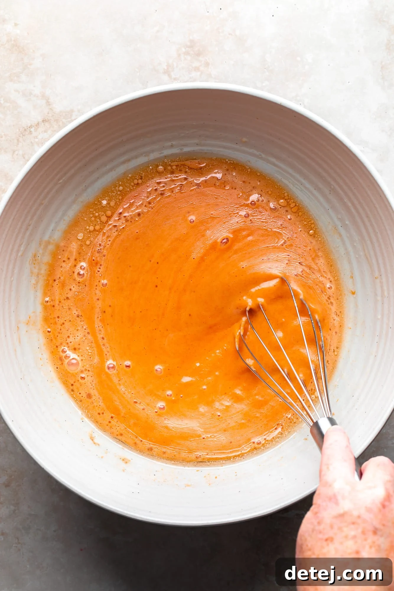 A woman's hand expertly whisking vegan pumpkin pancake batter in a large white bowl, ensuring a smooth consistency.