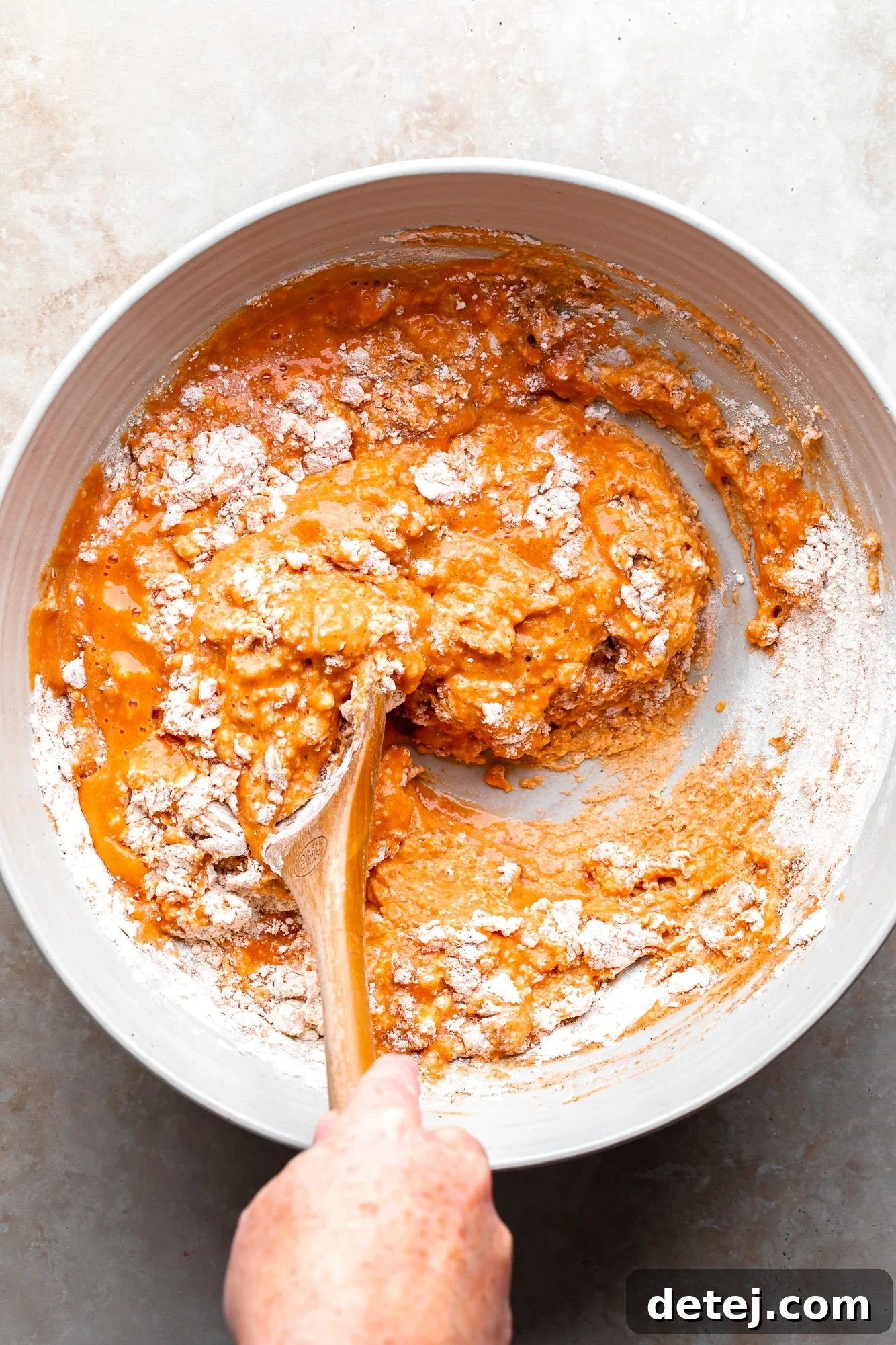 A woman's hand gently stirring vegan pumpkin pancake batter in a large white bowl, ensuring no overmixing for fluffy results.