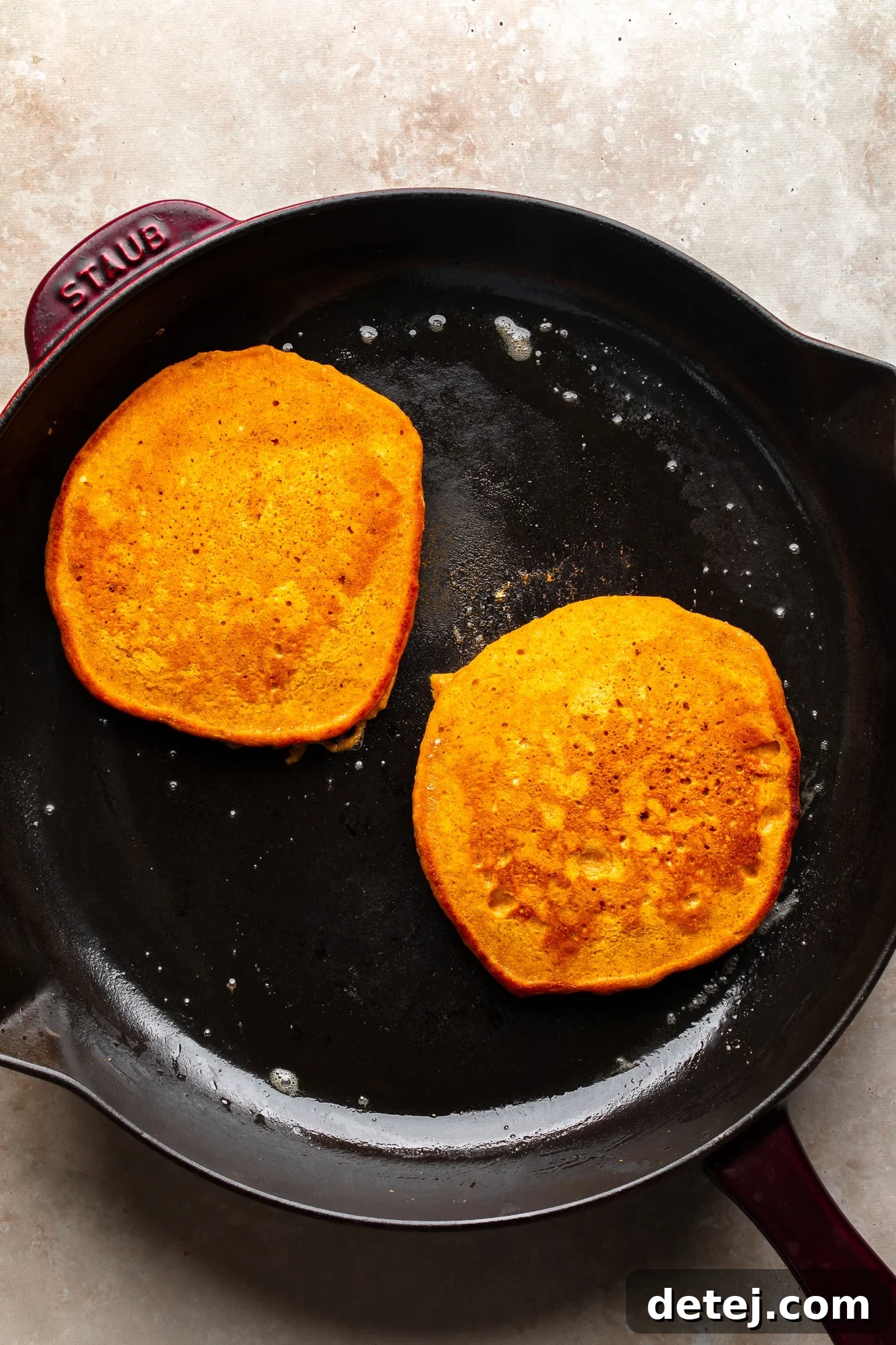 An overhead view of two vegan pumpkin pancakes sizzling and cooking to perfection in a black skillet, showing their readiness to be flipped.