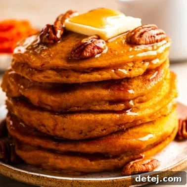 Close-up on a stack of vegan pumpkin pancakes, lavishly topped with flowing maple syrup, scattered pecans, and a pat of vegan butter.