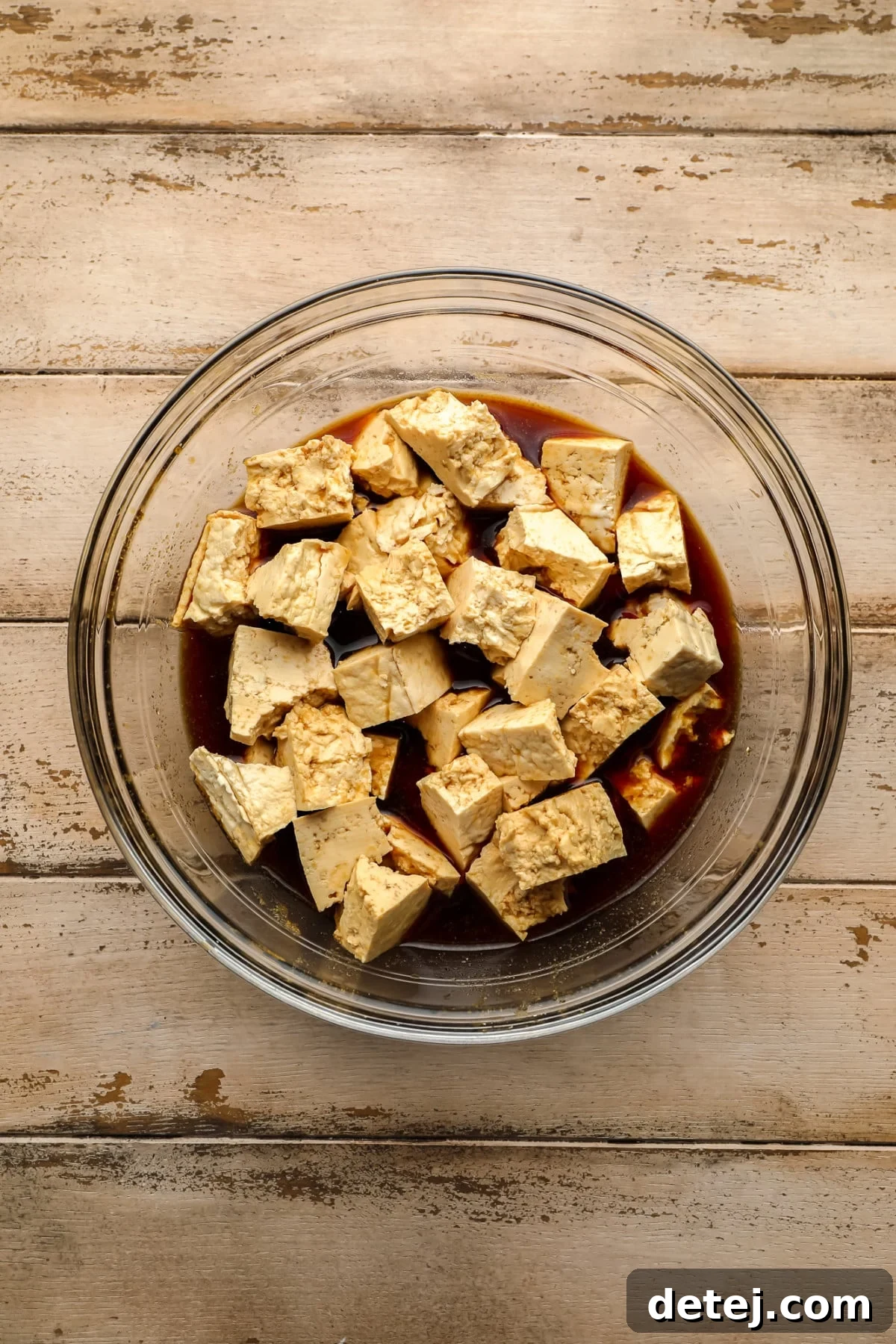 Crispy Plant Based Bites 4 Ripped tofu chunks soaking in a soy sauce marinade in a clear glass bowl, set against a rustic wooden background.