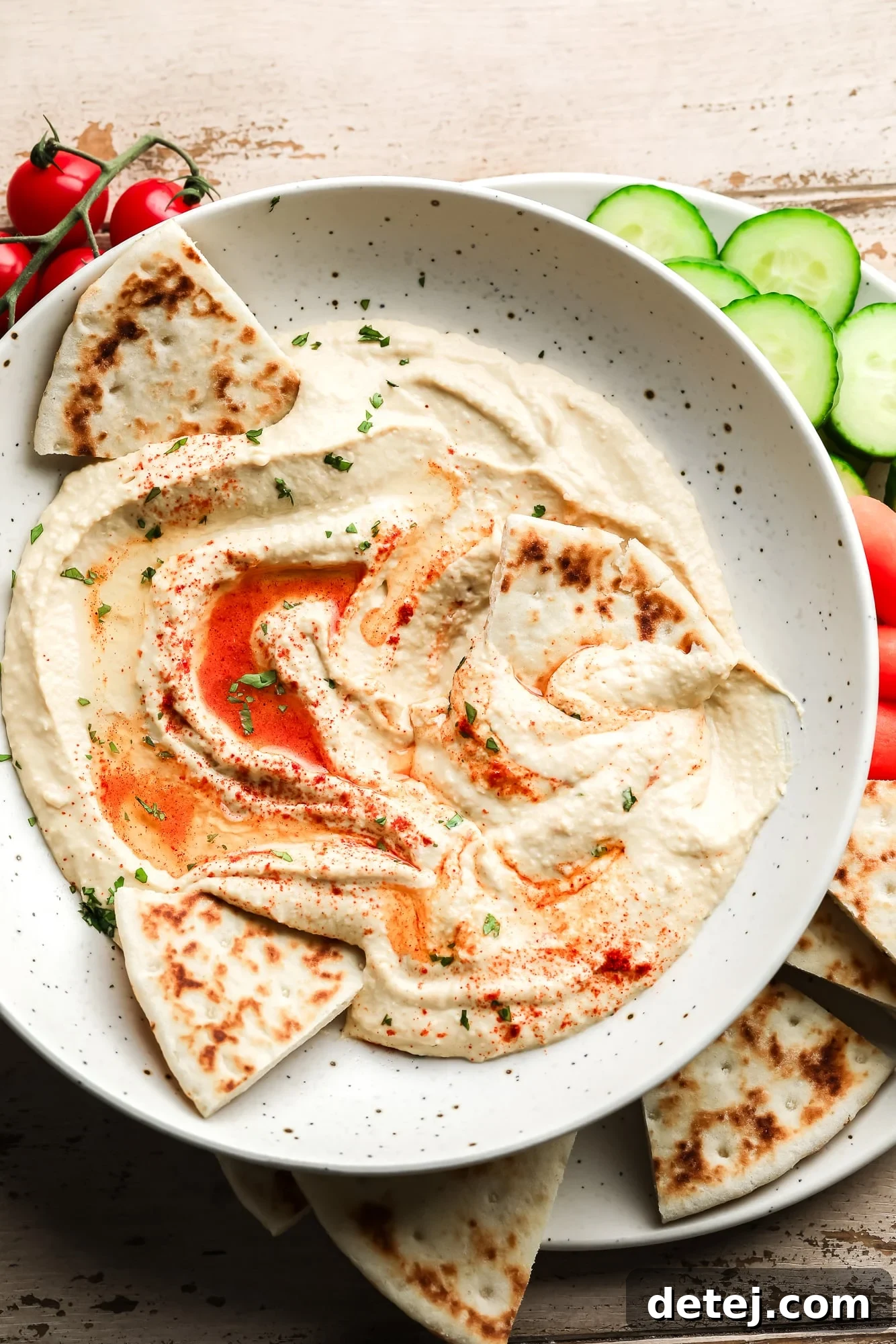 hummus topped with paprika and oil in a white bowl with pita bread and vegetables on the side.