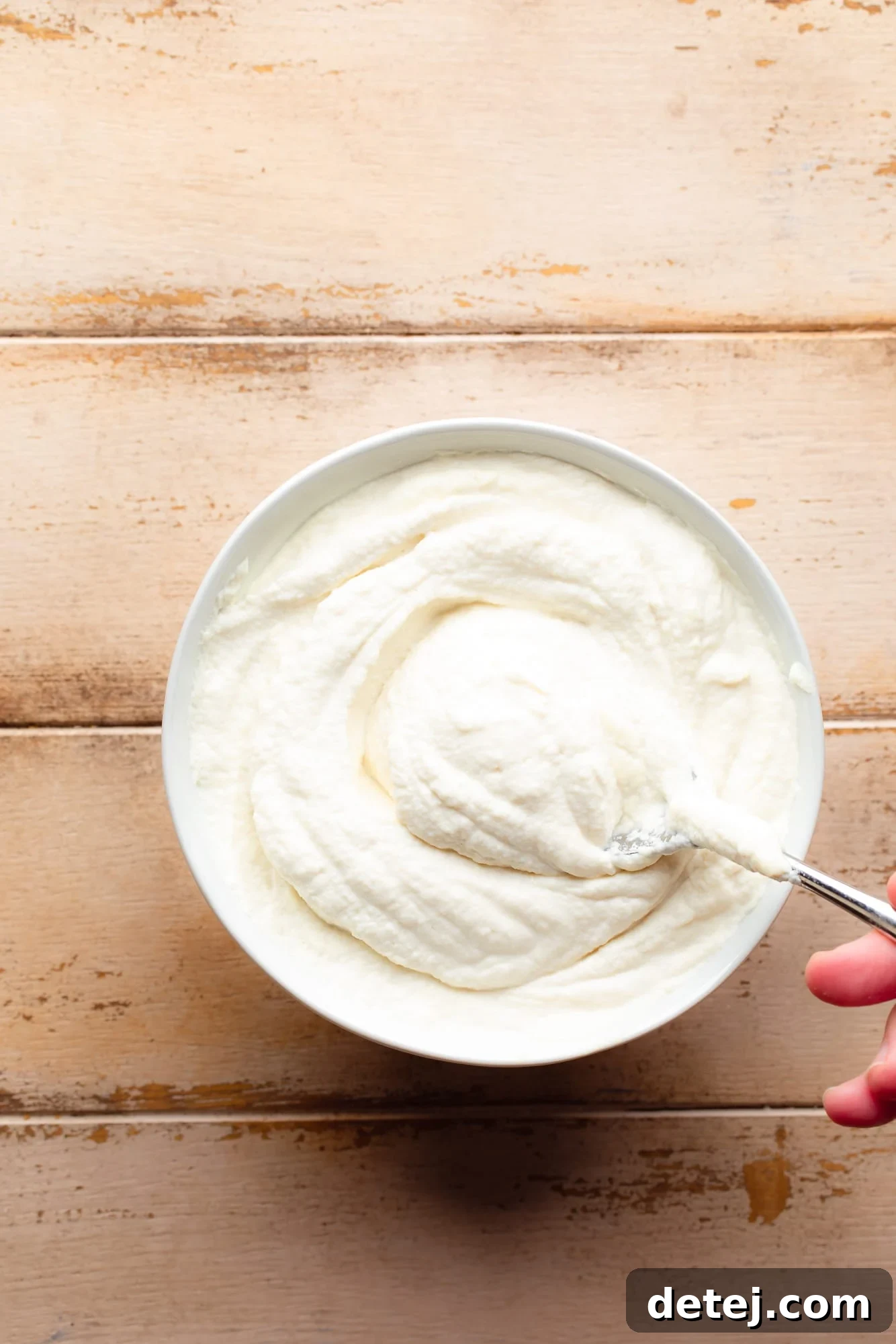A hand lifting a spoon out of a bowl filled with fluffy, homemade vegan ricotta cheese.
