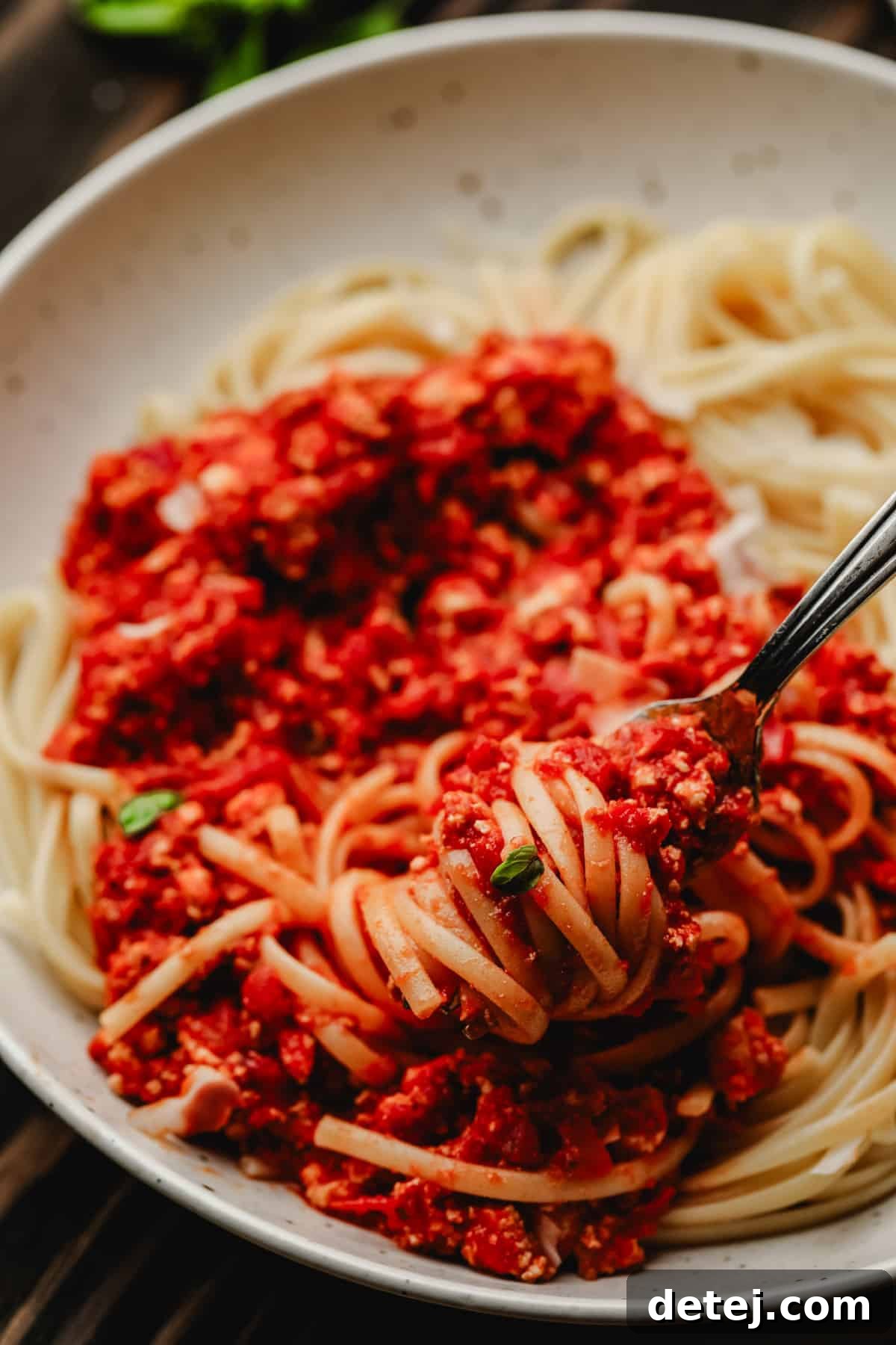 A speckled bowl filled with perfectly cooked pasta and rich, red vegan bolognese sauce, with a fork twirling a bite.