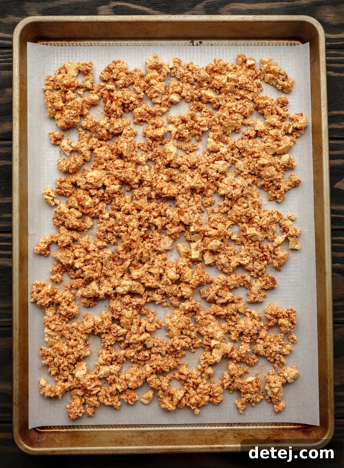 Uncooked, seasoned tofu crumbles spread in a single layer on a parchment-lined gold baking pan, ready for the oven.