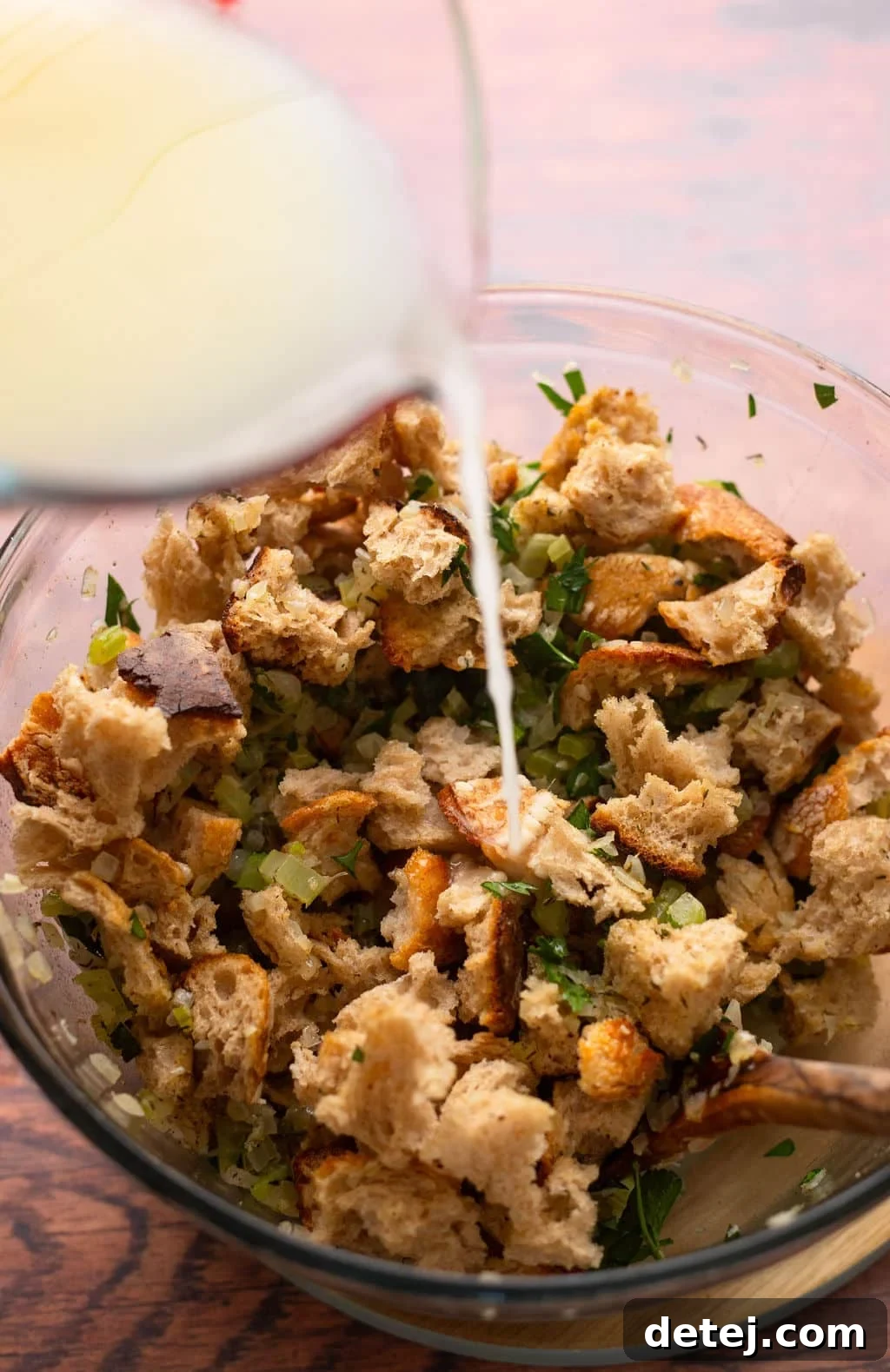 Ultimate Vegan Stuffing 8 Savory broth mixture being poured over bread cubes and vegetables in a mixing bowl.