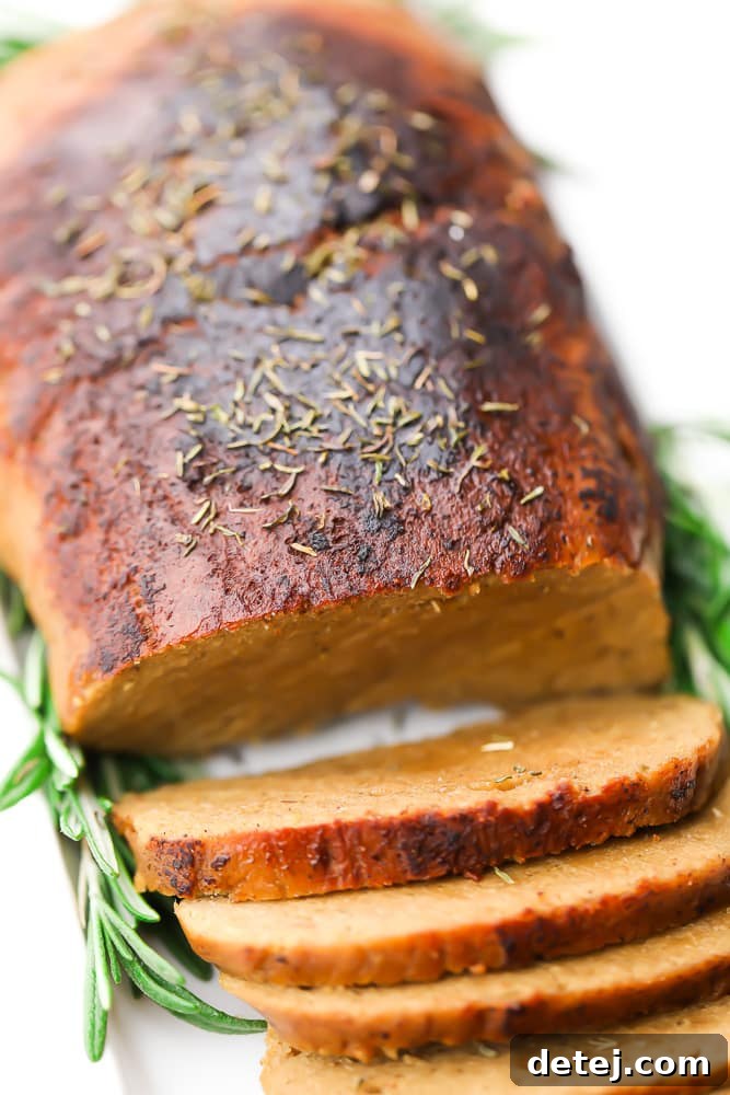 A vegan turkey loaf, perfectly browned with aromatic herbs, partially sliced on a pristine white plate, showcasing its succulent interior.