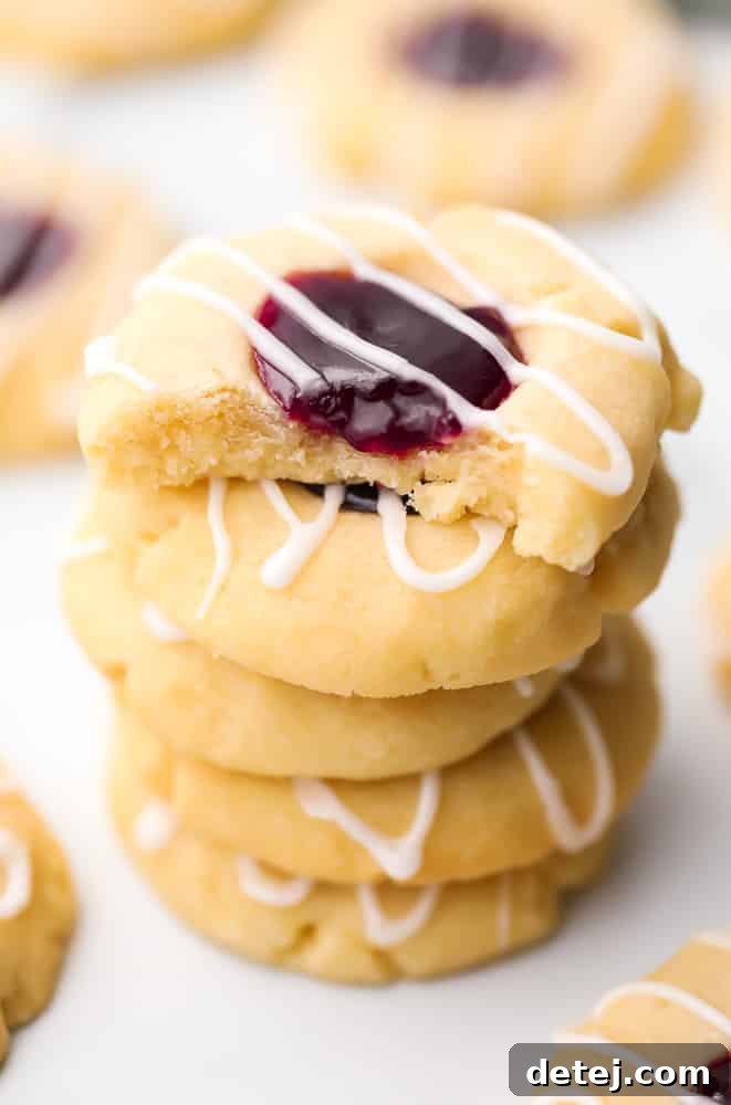 A vegan thumbprint cookie with a bite taken out of it, stacked on top of more cookies, showcasing the raspberry jam filling and golden-brown edges.
