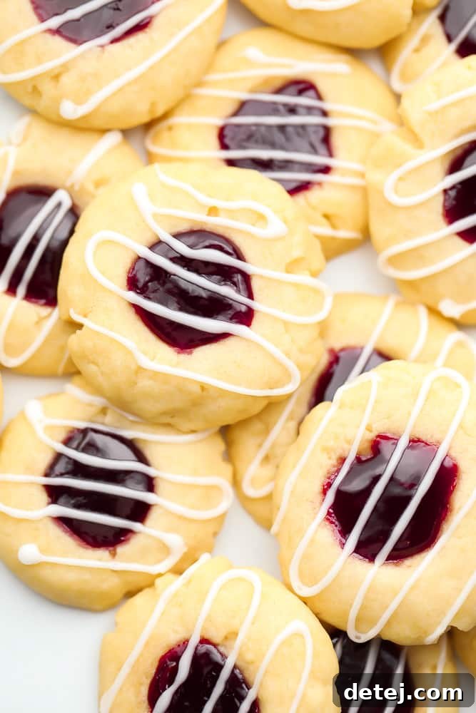 A close-up of numerous vegan thumbprint cookies filled with bright red raspberry jam and drizzled with a delicate white glaze, arranged beautifully.