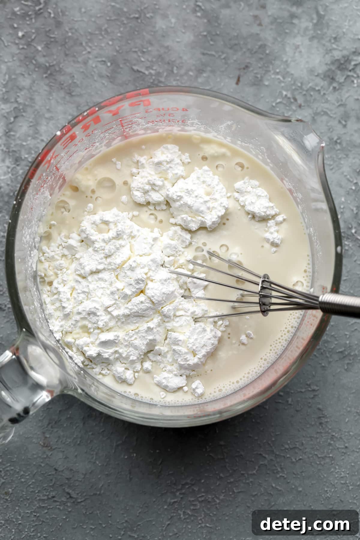 Whisking cornstarch into plant milk in a glass measuring cup, ensuring a smooth, lump-free mixture for the vegan custard.