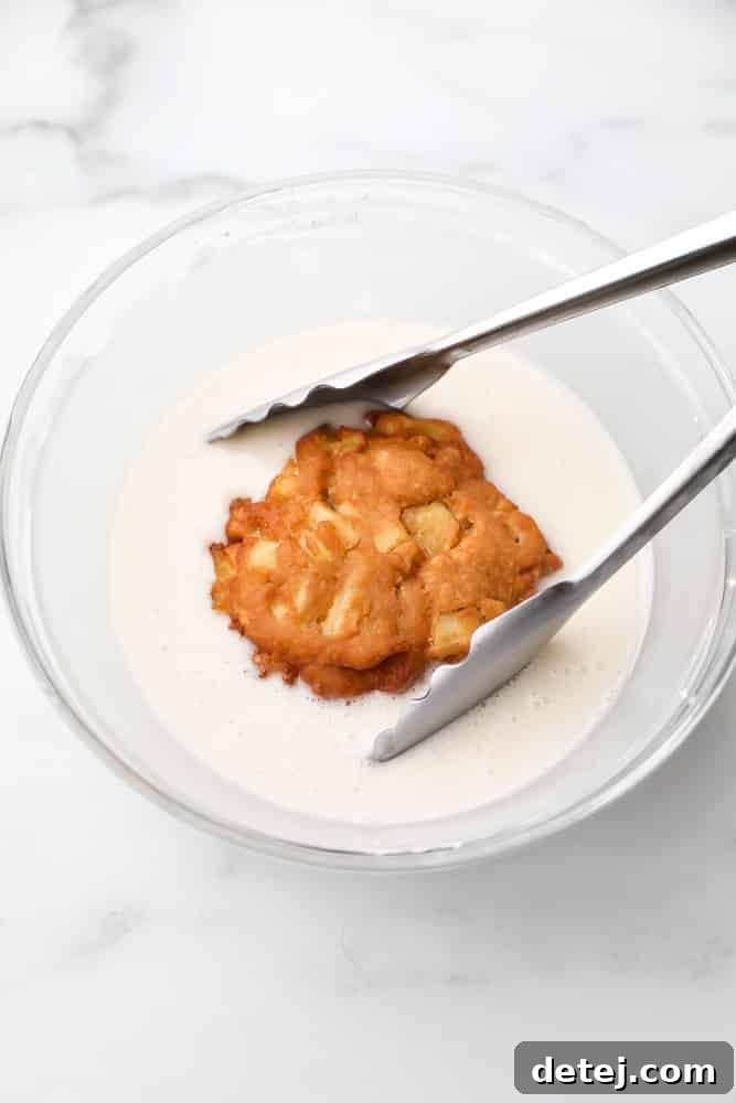 A hand dipping a freshly fried apple fritter into a bowl of sweet white glaze.