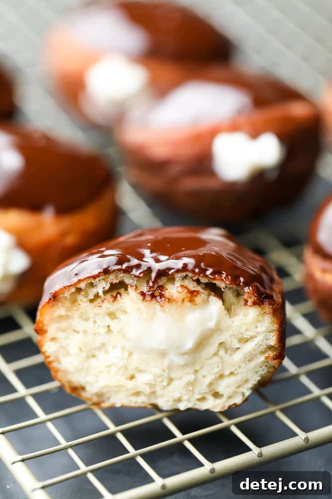 A close-up shot of a vegan Boston Cream Donut cut in half, revealing its creamy pastry filling, with more glazed donuts visible in the blurred background on a cooling rack.