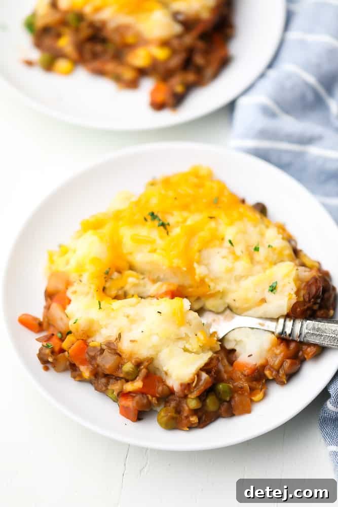 A close-up shot of a white plate holding a square slice of vegan lentil shepherd's pie, with a fork poised to take a bite, highlighting the golden-brown mashed potato topping and the rich, textured lentil filling beneath.