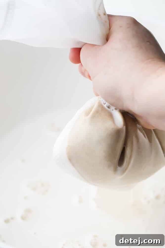 Hands gently squeezing a nut milk bag to extract creamy oat milk into a bowl.