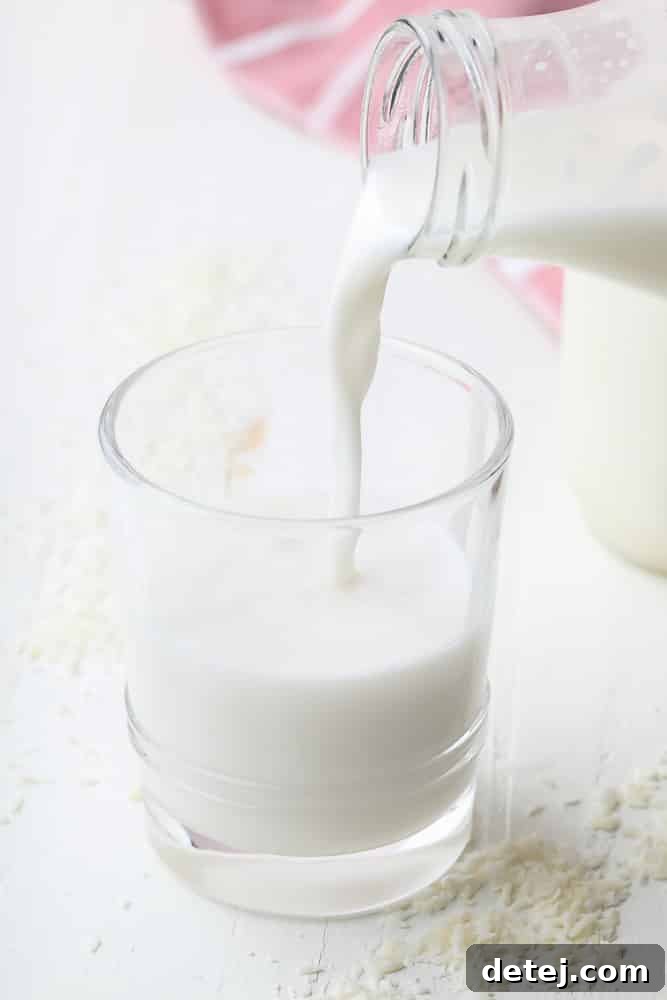 A glass of homemade coconut milk being poured from a jar, surrounded by fresh shredded coconut flakes, highlighting its creamy texture.