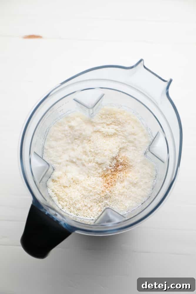 Unsweetened shredded coconut flakes, room temperature water, and a splash of vanilla extract gathered in a high-powered blender, ready for blending.