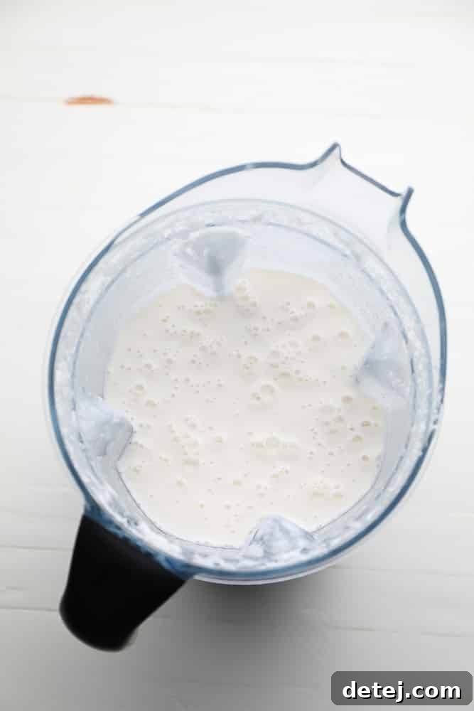 A high-powered blender filled with a frothy, creamy white mixture after blending shredded coconut and water, indicating readiness for straining.