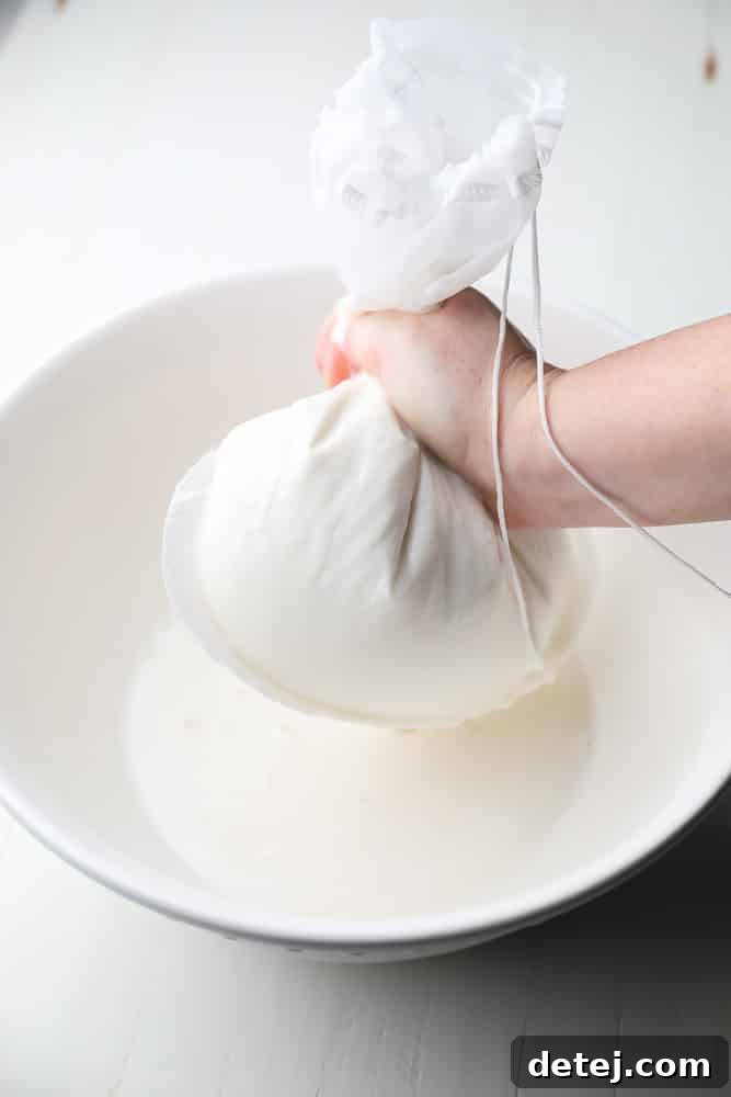 Homemade coconut milk being carefully squeezed through a white nut milk bag into a large glass bowl, separating the creamy liquid from the pulp.