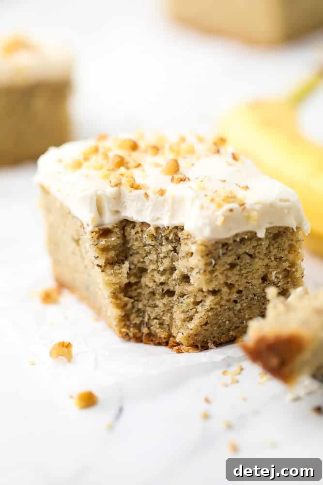 A beautifully frosted slice of vegan banana cake, featuring a soft, fluffy interior, topped with creamy vegan cream cheese frosting and a sprinkle of chopped walnuts. A fresh banana sits in the background.