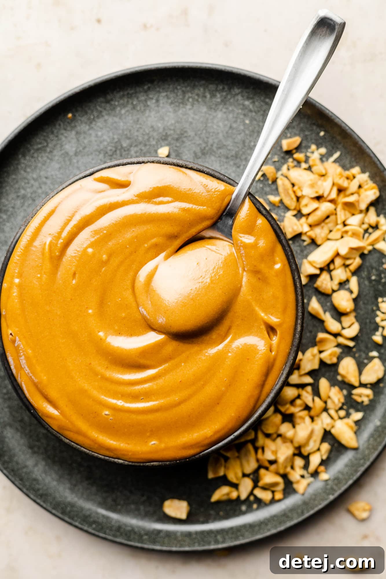 A spoon resting in a bowl of creamy peanut sauce, garnished with crushed peanuts on a sleek black plate.