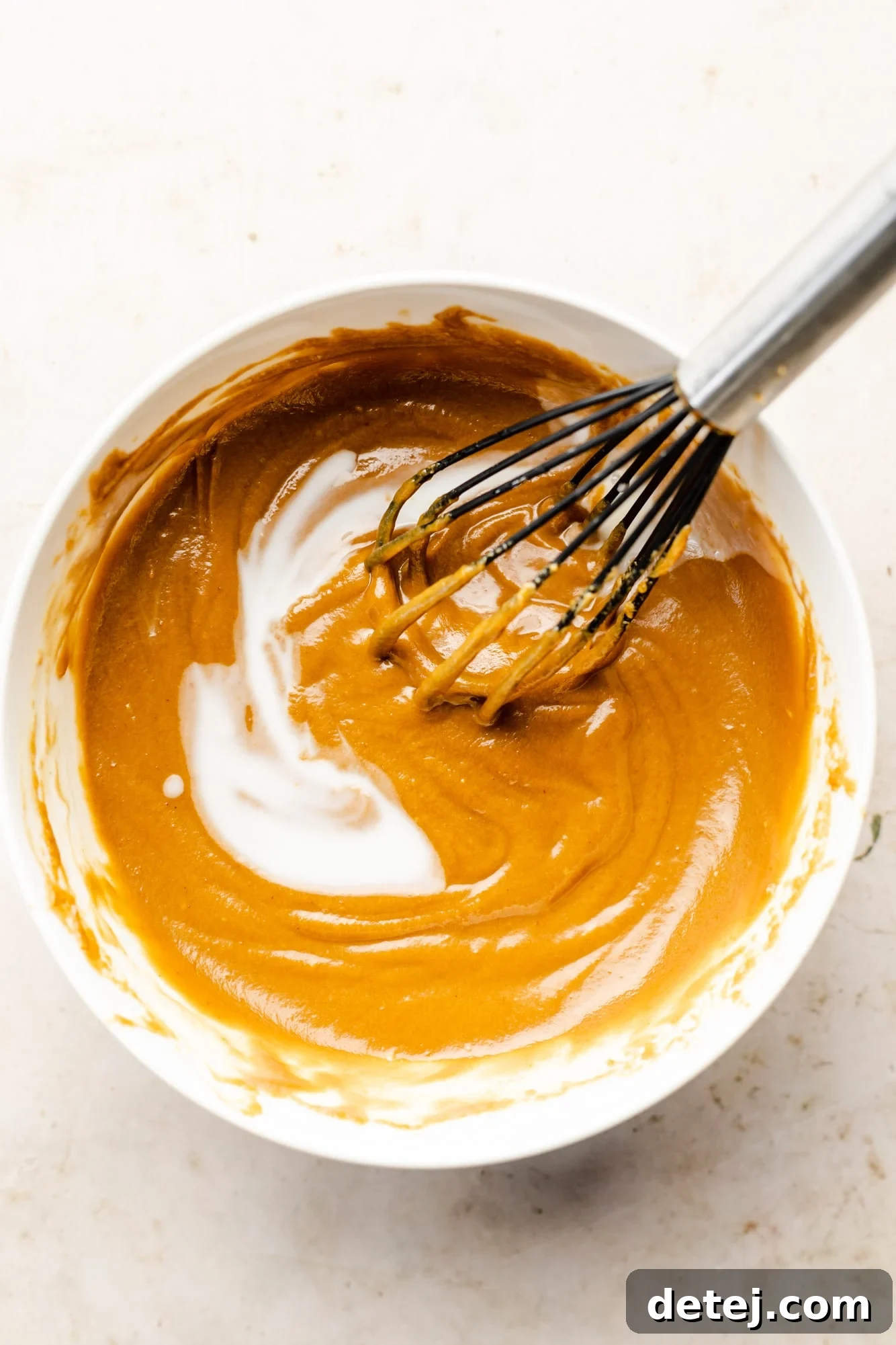 A hand using a whisk to incorporate coconut milk into a bowl of peanut sauce, making it smoother and creamier.