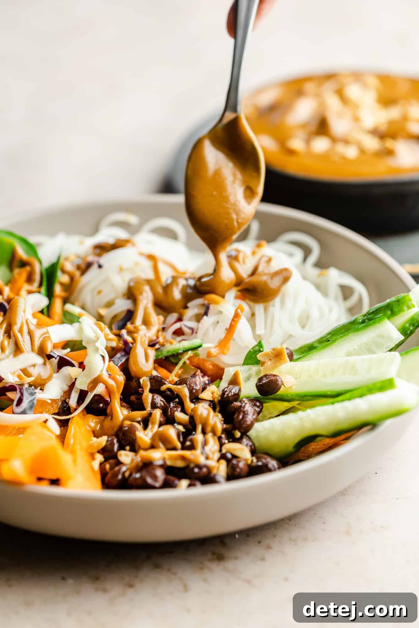 A spoon generously drizzling creamy peanut sauce over a colorful bowl of noodles and mixed vegetables, ready to be enjoyed.