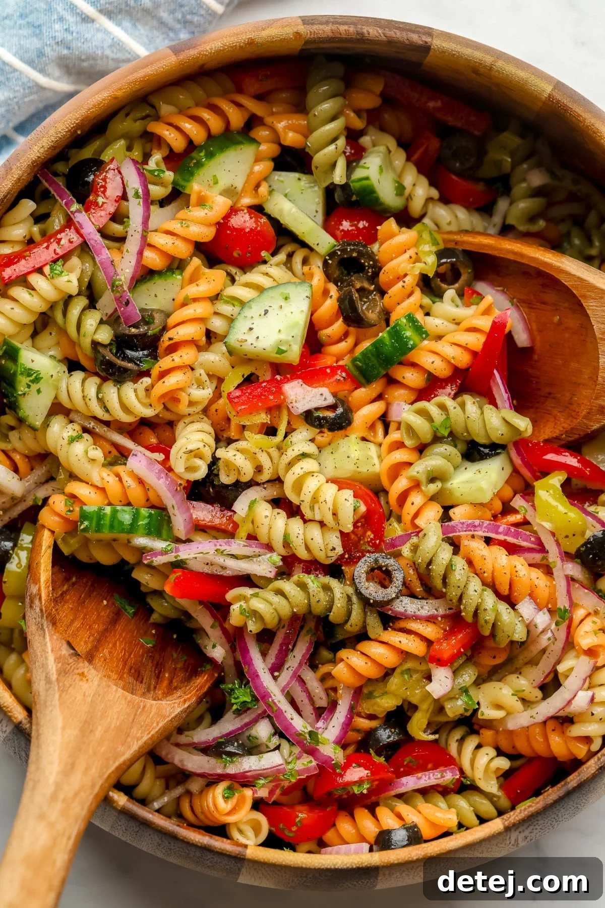 wood bowl with blue towel under it of vegan pasta salad with two wood spoons in it