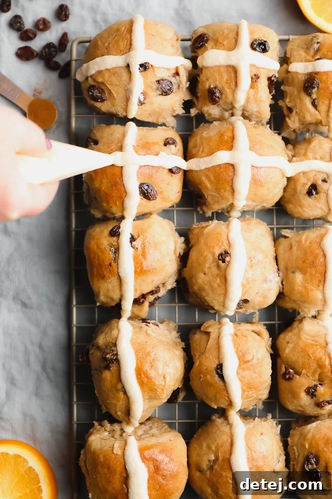 Freshly baked vegan hot cross buns being decorated with an orange glaze cross, showcasing their perfect golden-brown tops and inviting appearance.