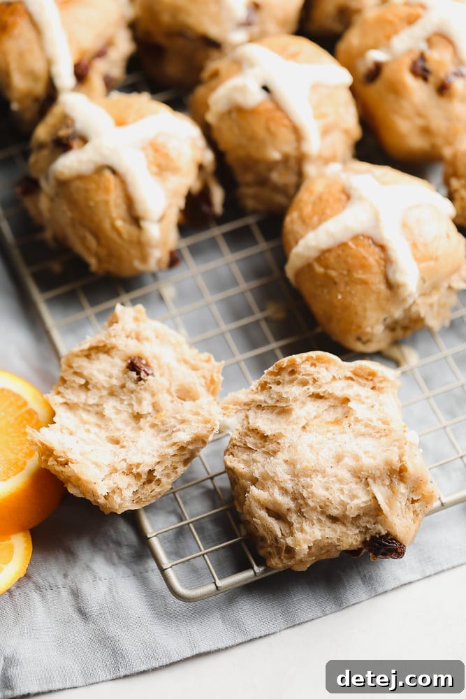 A freshly baked vegan hot cross bun, gently torn in half, revealing its incredibly soft, airy interior studded with juicy raisins and fragrant spices.