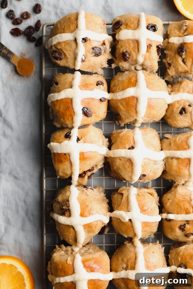 A batch of freshly baked vegan hot cross buns, glistening with a sweet sugar glaze and adorned with delicate orange icing crosses, cooling on a wire rack.