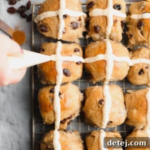 A square photo showing a baker piping cross shapes onto warm vegan hot cross buns, highlighting the decorative step.