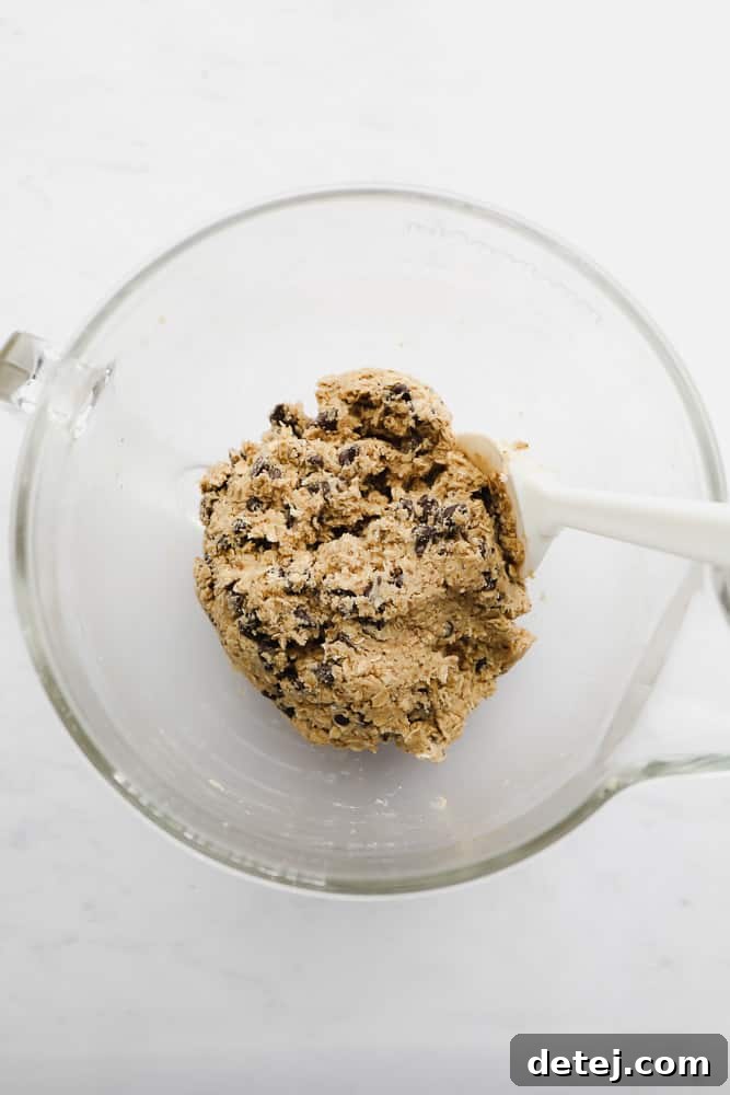 A close-up of raw vegan oatmeal chocolate chip cookie dough being stirred with a white spatula in a clear glass bowl, showing the texture of oats and chocolate chips.