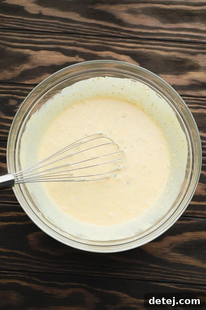 A close-up of a glass bowl holding a perfectly whisked, creamy yellow-tinted dressing with a whisk resting in it, against a warm wood background.