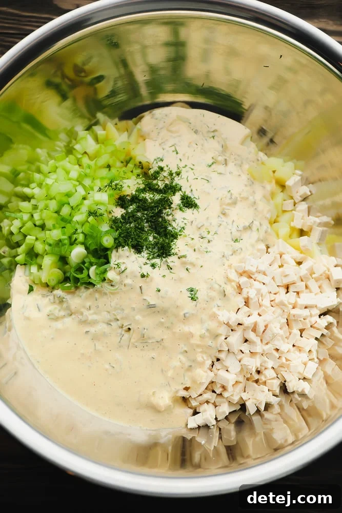 A silver mixing bowl on a wood background, brimming with warm, cooked potatoes, creamy sauce, diced celery, fresh herbs, and chopped tofu, all ready to be mixed into a delectable salad.