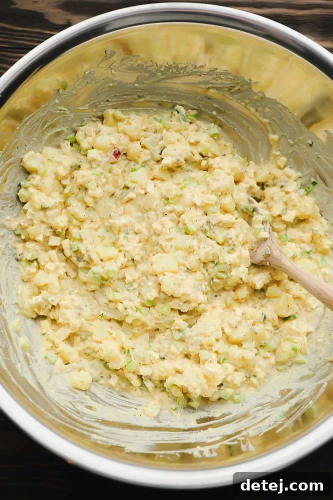 A silver bowl of vegan potato salad being stirred with a wooden spoon, showcasing its rich, creamy texture and hearty ingredients.