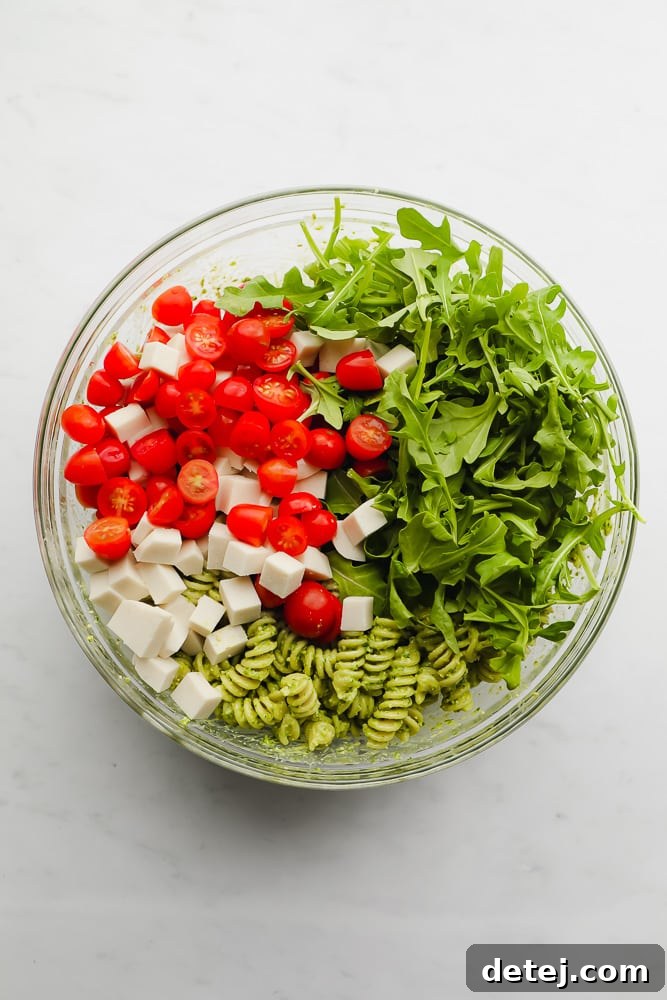 glass bowl filled with pesto pasta, arugula, cherry tomatoes, and cubed vegan cheese, ready for serving