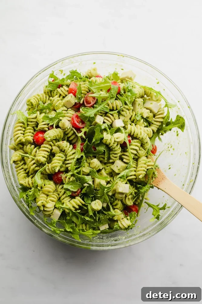 wooden spoon mixing a green pasta salad in a glass bowl, showcasing the fresh ingredients