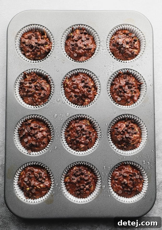 Unbaked vegan chocolate zucchini muffin batter, artfully sprinkled with extra chocolate chips, carefully portioned into paper-lined muffin cups within a baking tin, ready for the oven.