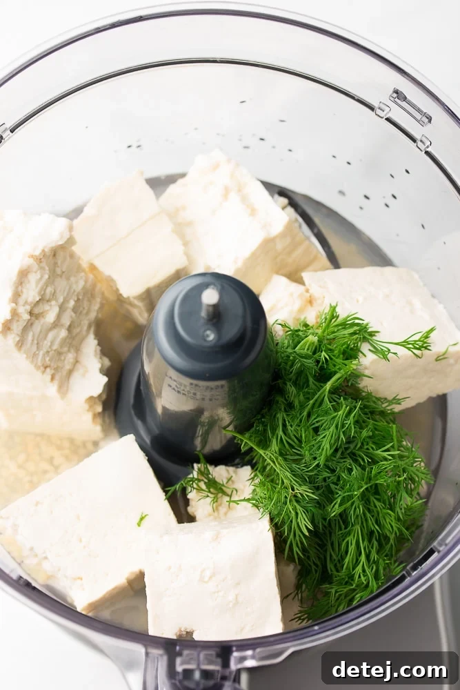 An overhead view of a food processor bowl, containing large chunks of firm tofu, fresh green dill sprigs, and garlic cloves, awaiting the blending process.