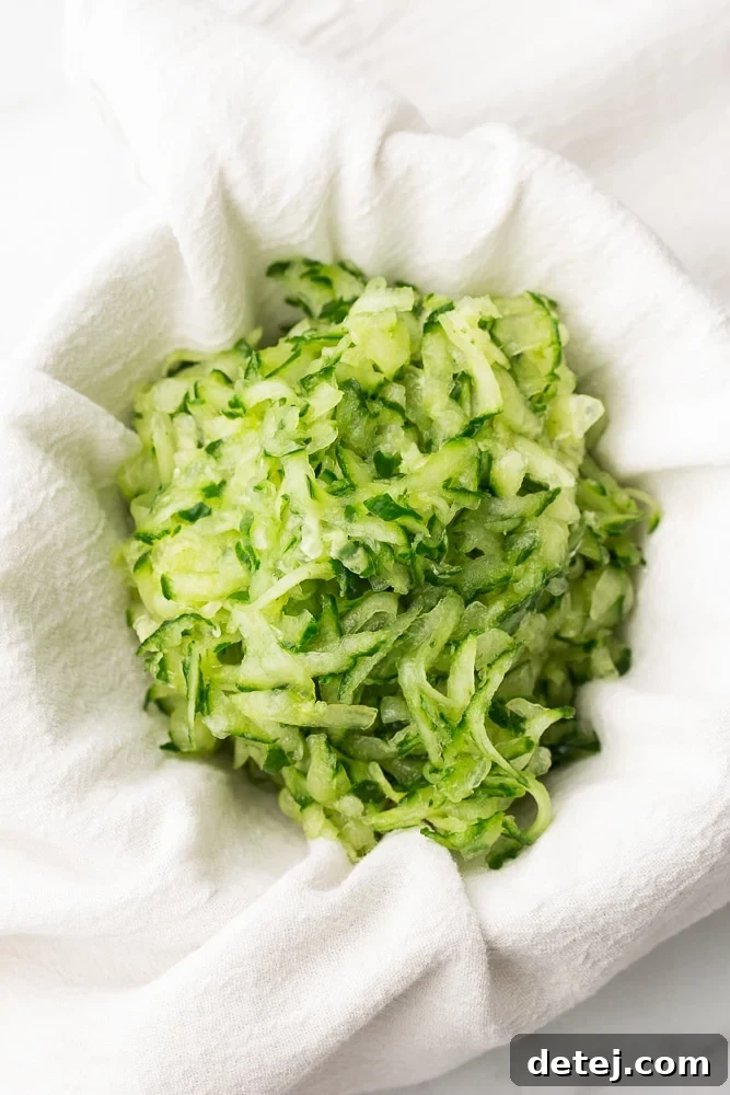 Close-up shot of grated cucumber being squeezed tightly within a white kitchen towel to remove excess moisture, with liquid dripping into a bowl below.
