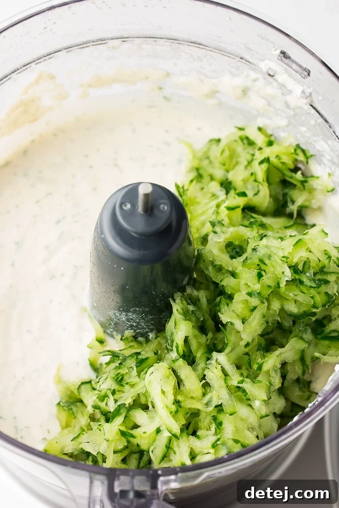 Overhead view of a food processor containing the creamy, blended tofu base with freshly grated cucumber added on top, before the final quick pulse.