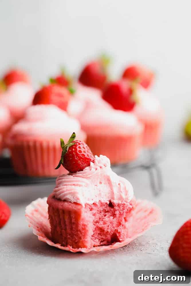 A vegan strawberry cupcake with a bite taken out, revealing the pink cake and strawberry pieces inside, topped with fluffy pink frosting.